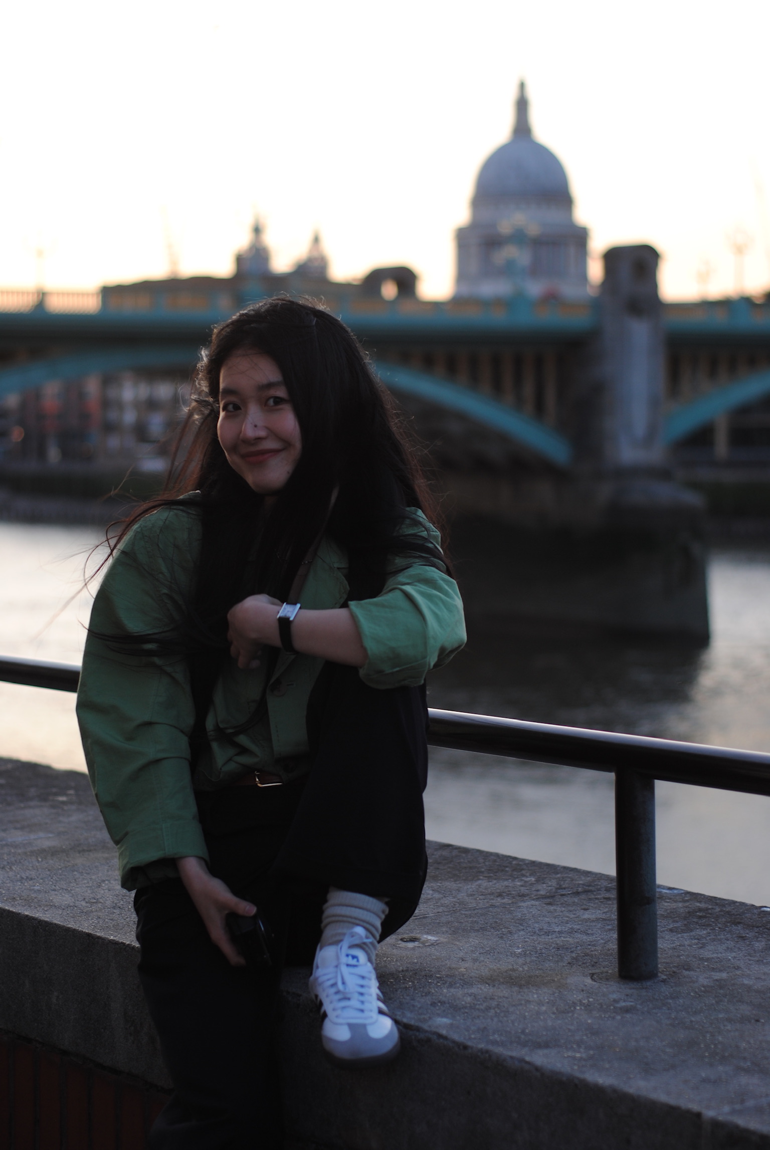 Woman sitting by riverside at sunset with bridge silhouette